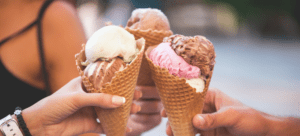 A trio of hands raising ice cream cones in a toast, featuring scoops of chocolate, vanilla, and pink strawberry ice cream