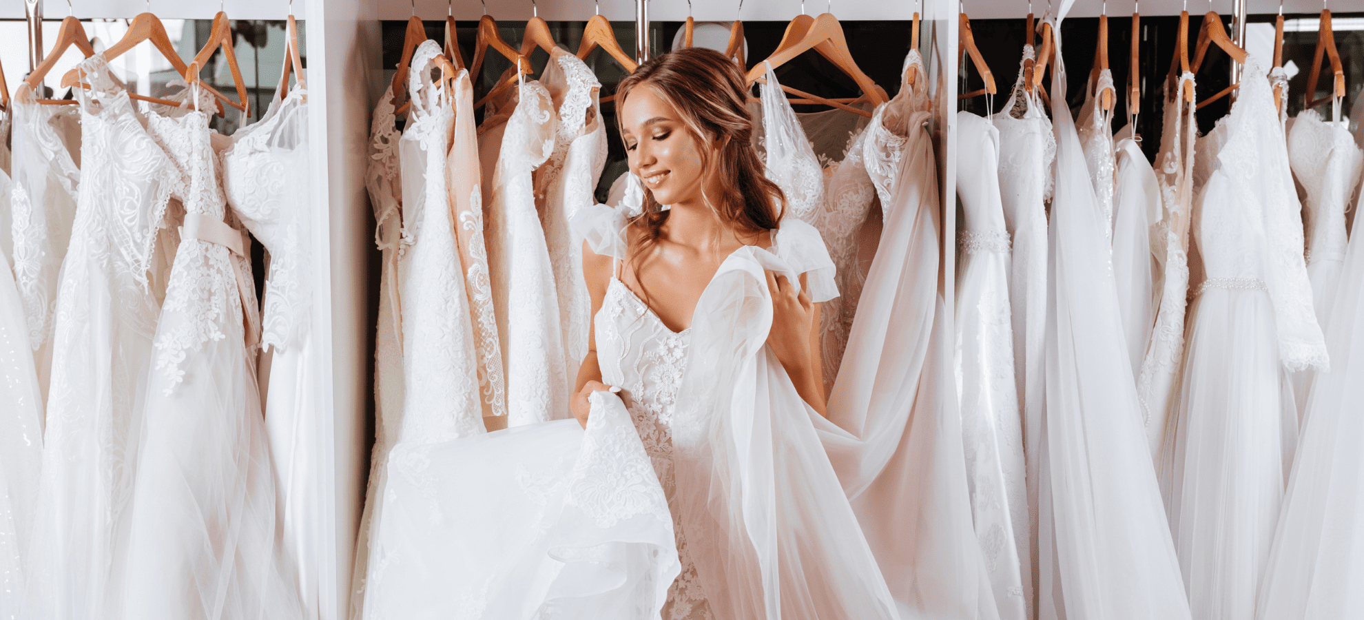 A woman stands in front of a rack filled with white wedding dresses at one of the Myrtle Beach bridal boutiques, holding up one of the dresses and smiling. The dresses have various lace patterns and designs, and the woman looks happy and elegantly dressed in a white lace gown.