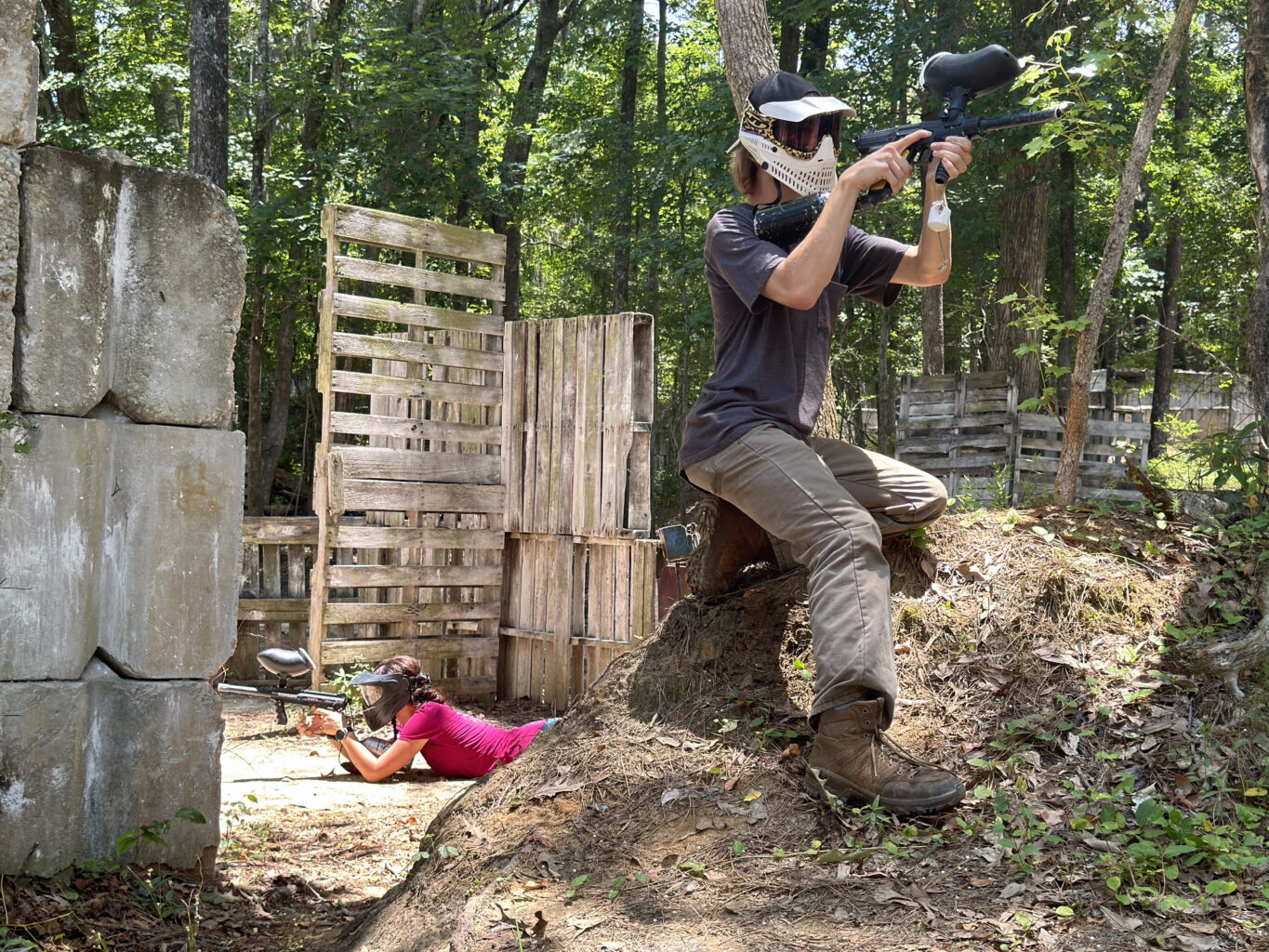 man posing with a paintball gun and woman laying on the floor holding a paintball gun in a field of obstacles.