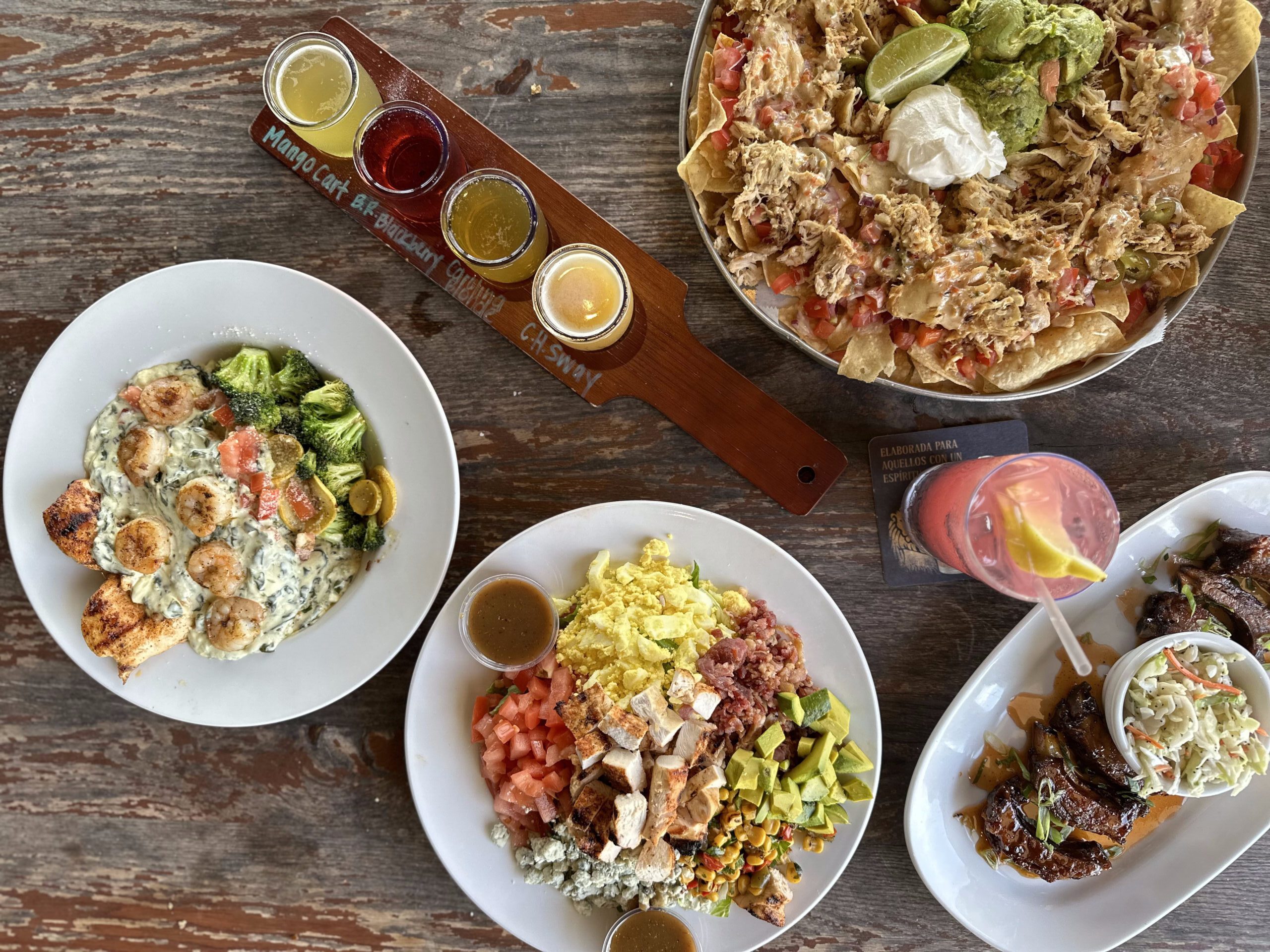 Aerial shot of a table displaying diverse meals. Left: grilled shrimp, broccoli, and creamy spinach sauce. Center: a chopped salad featuring scrambled eggs, avocado, diced tomatoes, and grilled chicken. Right: nachos topped with shredded chicken, sour cream, and salsa. Drinks include three craft beers and a pink cocktail with a lemon slice.