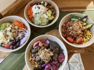 four salad bowls with different condiments ranging from corn, olives, red onions, cherry tomatoes, cucumbers, cheese and others